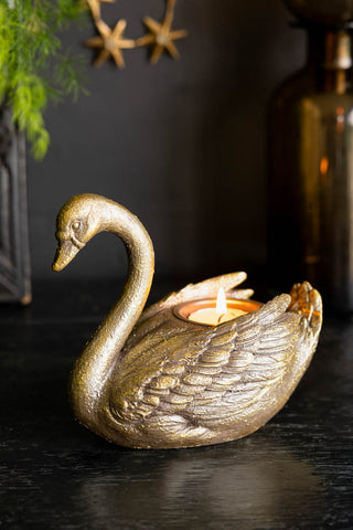 The Gold Swan Tealight Holder shown against a black wall with gold accessories. 