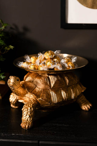 The Gold Tortoise With Tray displayed with wrapped chocolates on, styled on a dark sideboard with a plant and an art print.