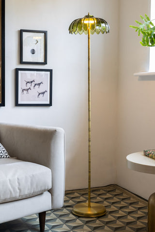 The Gold & Green Desert Palm Floor Lamp switched on and displayed in the corner of a room next to a sofa and a side table.