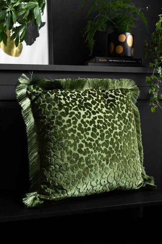 The Green Leopard Velvet Cushion with Fringe Trim styled on a black bench with plants and accessories in the background.