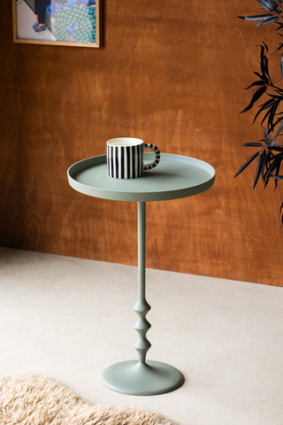 Lifestyle image of the Anjou Metal Side Table - Sage Green styled in front of a dark wooden wall, with a striped mug, art print, plant and rug. 