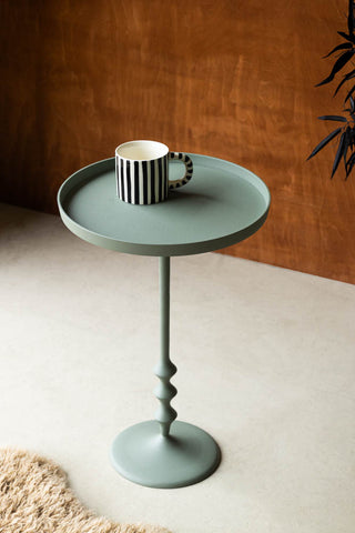 Lifestyle image of the Anjou Metal Side Table - Sage Green styled in front of a dark wooden wall, with a striped mug, plant and rug.