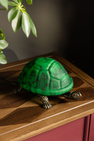 The Green Speckled Glass Turtle Table Lamp shown on a wood tabletop against dark walls. 