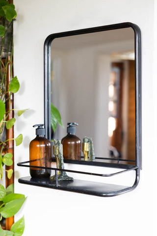 Lifestyle image of the Gun Metal Mirror With Shelf styled on a white wall with an amber bottle and candle next to a plant.