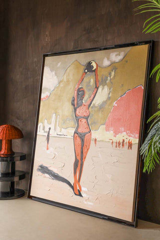 Lifestyle image of the HKliving Playground Framed Artwork displayed leaning against a dark wood wall, styled with a side table, table lamp and plant. 