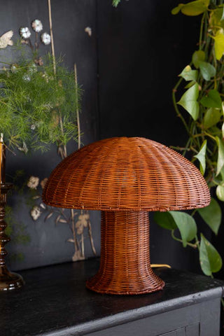 Lifestyle image of the HKliving Rattan Toadstool Table Lamp styled on a black sideboard with greenery and home accessories. 