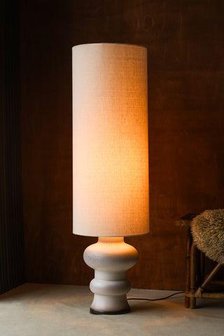Lifestyle image of the HKliving White/Brown Retro Reactive Glaze Lamp displayed illuminated in front of a dark wooden wall next to a wicker bench with fluffy rug on.