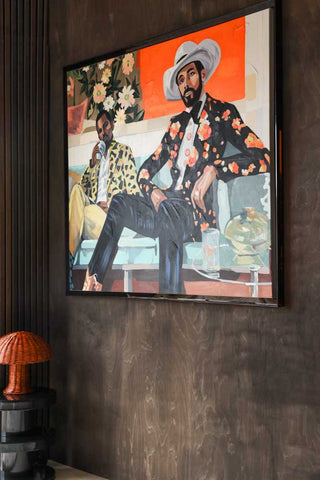Lifestyle image of the HKliving Saloon Framed Artwork displayed on a dark wood wall and styled with a side table and table lamp. 