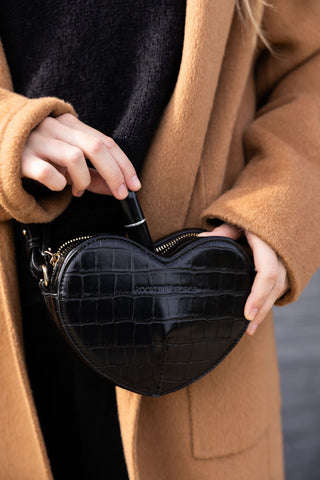 A black heart shaped bag being held by a model, placing a lipstick inside.