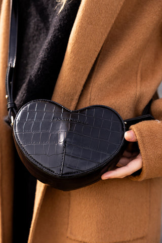 A black heart shaped bag being worn across a model's body and being held in their hand.