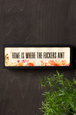 The Home Is Where The Fuckers Aint Light Box switched on and displayed on a dark wall above a plant.