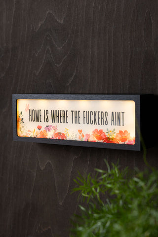 The Home Is Where The Fuckers Aint Light Box switched on and displayed on the wall above a plant.