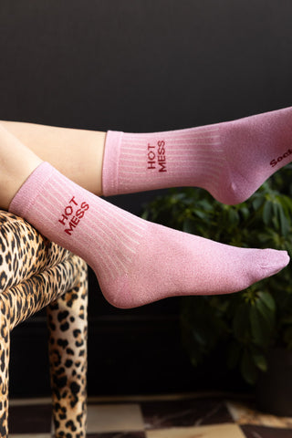 The Hot Mess Pink Glitter Socks shown on a model hanging off the edge of a leopard print bench against a black wall. 
