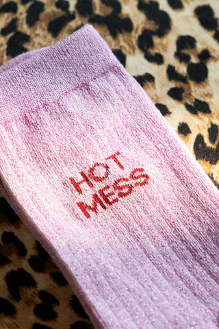 Close up detail of the Hot Mess Pink Glitter Socks against a leopard background. Socks are pink with burgundy writing that says 'hot mess'.