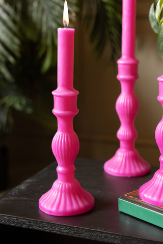 Hot Pink Candlestick Shaped Candle - Medium shown with the candle lit, with plants and the tall version of the candle in the background of the shot. 