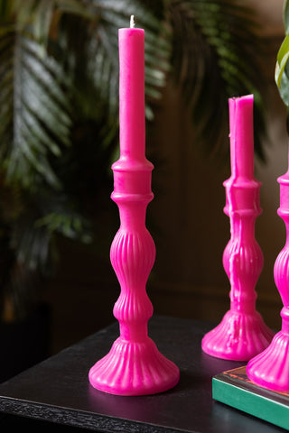 The Hot Pink Candlestick Shaped Candle - Tall shown unlit against a background of plants. 