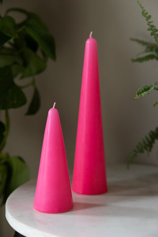 The Small and Large Hot Pink Cone Shaped Candles displayed together on a white table with some plants in the background.