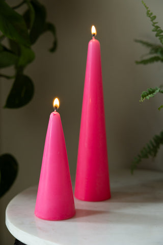 The Small and Large Hot Pink Cone Shaped Candles displayed together lit on a white marble table with some plants in the background. 