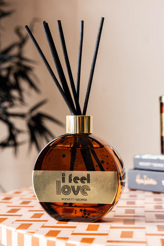 The I Feel Love Cedarwood & Frankincense Reed Diffuser displayed on a patterned surface with a plant and some accessories in the background.