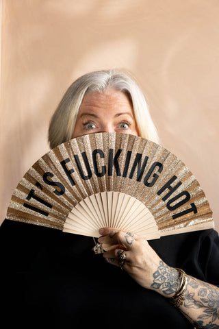 The It's Fucking Hot Gold Glitter Fan shown being held up against a face with a neutral background. 