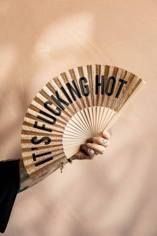 The It's Fucking Hot Gold Glitter Fan shown against plaster walls. 