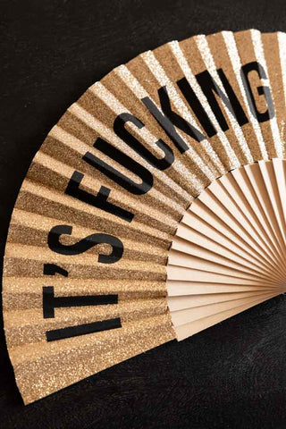 The It's Fucking Hot Gold Glitter Fan shown flat on a black table. 