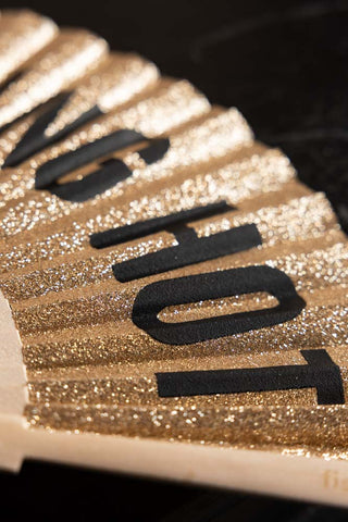 A detail showing the gold glitter and text detailing on the It's Fucking Hot Gold Glitter Fan. 