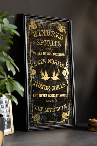 Image of Kindred Spirits Decorative Mirror against a black wall with a plant in the corner. 