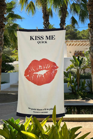 The Kiss Me Beach Towel hanging in the sunshine surrounded by trees and plants.