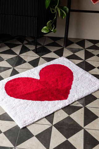 The LOVE Tufted Cotton Bath Mat on a black and white geometric floor, with a black cupboard, towel rack, towel and plant in the background.