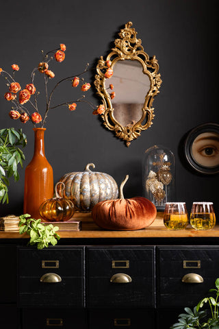 A lifestyle image with a Halloween theme, showing the Large Rustic Patina Glass Pumpkin amongst other pumpkins, an apothecary sideboard and a gold ornate mirror against a black wall. 