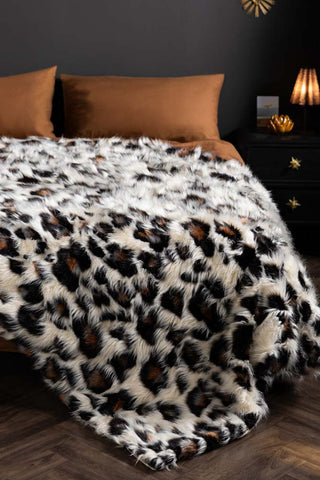 The Leopard Faux Fur Throw show with tan bedding in a bedroom with black walls and a black bedside table. 