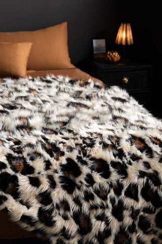 The Leopard Faux Fur Throw shown on a bed with tan bedding and a bedside table in the background. 