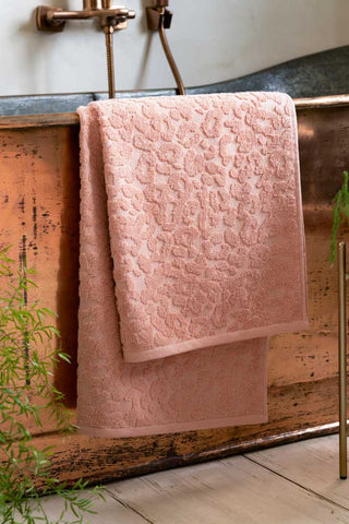 A Leopard Jacquard Towel in Dusty Pink shown hanging from the side of a copper bath tub. 