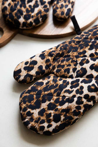 Close-up of the Leopard Love Print Oven Mitts on a work surface with a wooden chopping board.