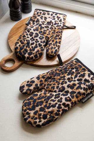 The Leopard Love Print Oven Mitts displayed on a kitchen worktop with a wooden chopping board and some salt and pepper grinders.