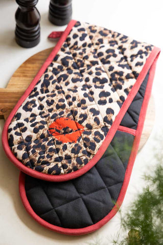 The Leopard Love Print & Red Detailing Oven Gloves displayed on a wooden chopping board with some salt and pepper grinders in the background.