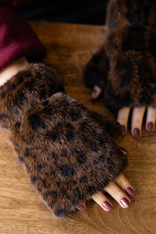 The Leopard Print Faux Fur Handwarmers being worn by a model, resting their hands on a wooden surface.