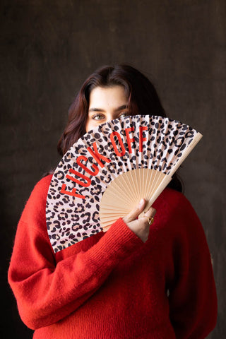 The Leopard Print 'Fuck Off' Wooden Hand Held Fan being held by a model in front of her face.