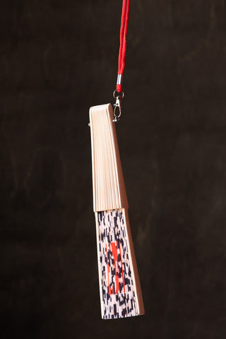 The Leopard Print 'Fuck Off' Wooden Hand Held Fan folded up and hanging from its attached loop, in front of a dark background.