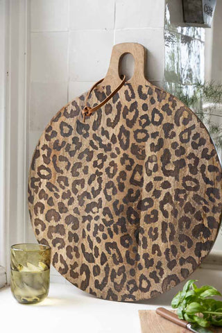 Lifestyle image of the Lifestyle image of the Leopard Print Mango Wood Serving Board displayed in front of a tiled kitchen wall and styled with a plant and various kitchen accessories. 