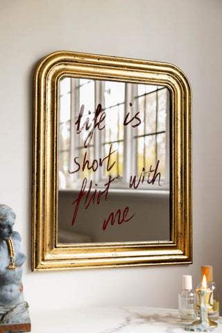 The Life Is Short, Flirt With Me Mirror shown against white walls with candles and accessories in the background. 