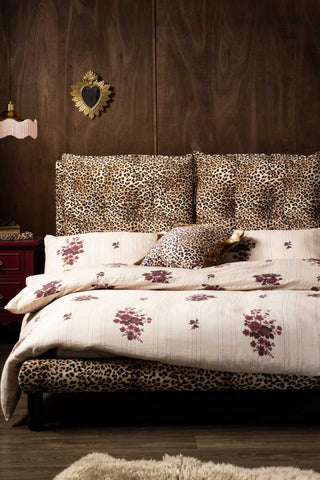 The Lounge Lover Bed in Leopard Print Velvet shown head on with pink floral bed linen against a dark textured wood wall. 