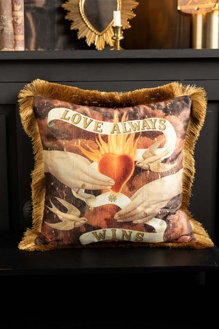 The Love Always Wins Fringed Velvet Cushion styled on a black seat in front of a black cabinet with accessories on.