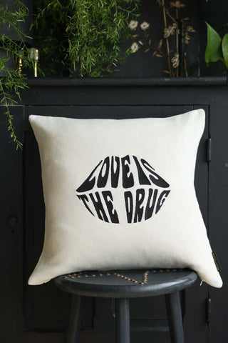 The Love Is The Drug Knitted Lips Cushion displayed on a black stool in front of a cabinet, with plants and home accessories in the background.