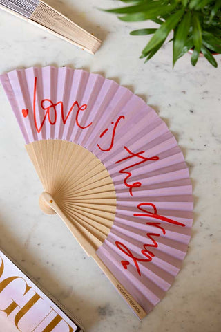The Love Is The Drug Pink & Red Fan displayed on a white marble table with another fan, magazine and a plant.