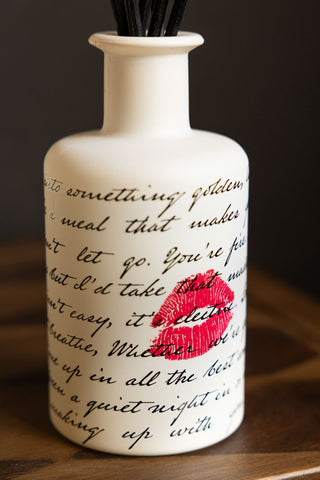 The design of the Love Letters Reed Diffuser, featuring love letter quotes in black text and a red kiss against the white diffuser. set includes black reeds. 