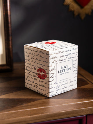 The box of the Love Letters Scented Candle, with the same love letter notes and red kiss marks, shown on a wooden console against black walls. 