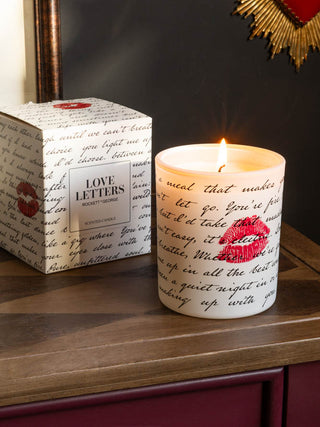 The Love Letters Scented Candle shown with the box in the background on a wooden console table, against dark black walls. 