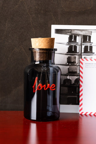The Love Neon Bottle Light on a wooden surface with some postcards leaning on the wall behind.